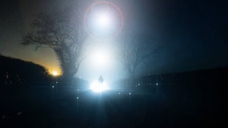 A supernatural, UFO concept of a scary, mysterious hooded figure, standing in front of a bright lights in the sky, on a country lane, on a spooky foggy winters night.の写真素材
