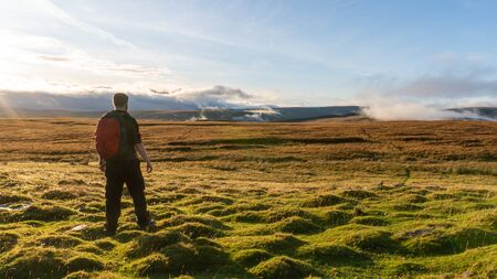 A male hiker looking out across a bleak moorland as the sun sets. With clouds covering hills in the...