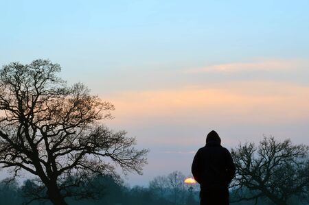 A silhouette of a hooded figure standing in a field, watching the sunset on a winters afternoon. の写真素材
