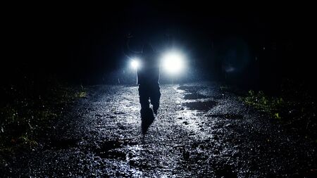 A moody hooded figure at night kneeling in front of car headlights with his hand up. As heavy rain comes down.の写真素材