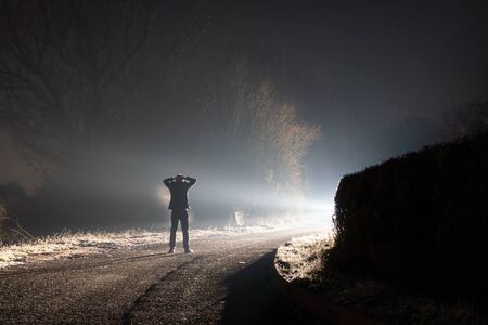A figure standing on a road surrendering with his hands on his head. Looking at a bright light.  On a spooky country lane. On a misty winters night.の写真素材