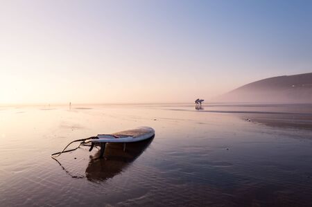 A surfboard on a beach relflected by the water on the sand. On a beautifully misty afternoon....