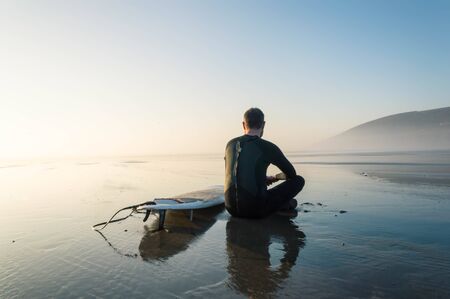 A surfer sitting by a surfboard on a misty afternoon looking out tosea. Saunton, Devon, UK. の写真素材