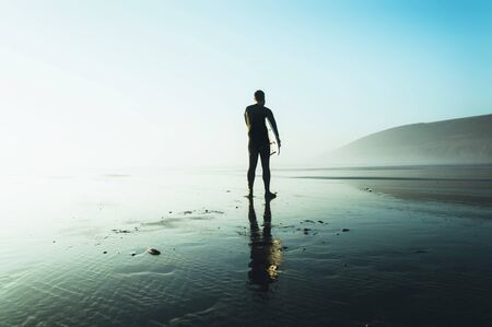 A surfer holding a surfboard silhouetted against the afternoon sun, on a misty winters day. Saunton,...