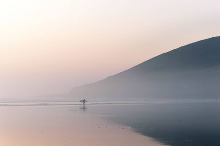 A lone surfer walking along the beach on a misty winters afternoon. Saunton, Devon, UK.