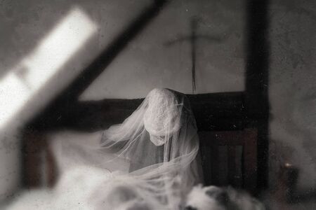 A ghost like figure, sitting on a bed in a corner of an old timber framed building. With a blurred,...