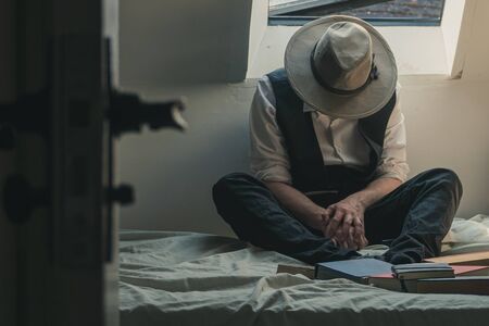 A man looking down wearing a fedora hat and waistcoat sitting on a bed with books surrounding him.の写真素材