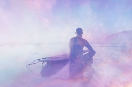 Silhouette of a surfer with surfboard, sitting on a beach looking at the sea. With a double exposure of the sky at sunset and an abstract, experimental dream like edit.の写真素材