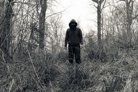 A scary, hooded figure with glowing evil eyes and blank black space where his face should be, standing in a forest in winter. With a vintage edit.の写真素材