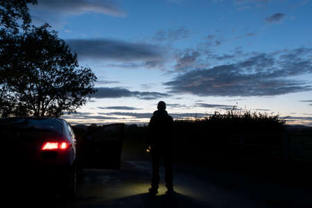 A man next to a car, standing in the middle of a road, silhouetted against the evening  sky,