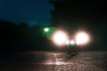 A man in front of a car, standing in the middle of a road, silhouetted against car headlights