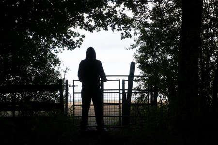 A hooded figure standing by a gate on the edge of a forest, silhouetted against the sky.の写真素材