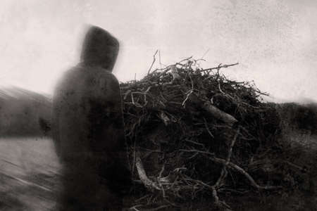 A spooky, demonic, hooded figure, standing next to a pile of branches in the countryside. With a...