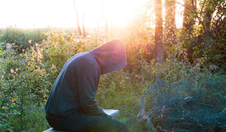 A mental health concept of a moody, hooded figure, sitting on a bench. Back lighted by the evening sun.の写真素材