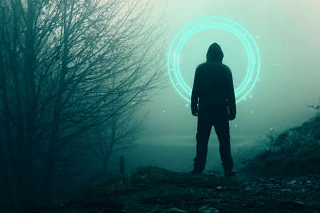 A spooky hooded figure, back to camera. Looking at a glowing, portal, gateway. In a forest on a foggy, winters day.の写真素材