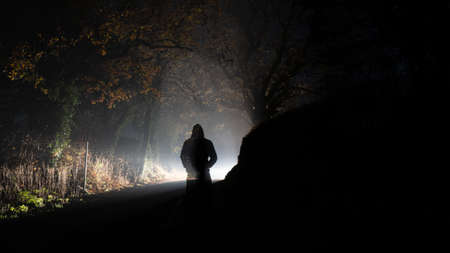 A ghostly transparent hooded figure silhouetted against a glowing bright light on a country road. On a foggy winters night.の写真素材