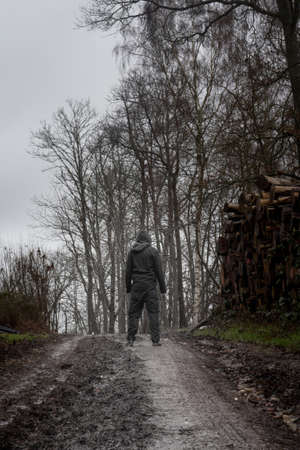 A hooded man, back to camera on a muddy path, next to a log pile on a grey moody day in the countrysideの写真素材
