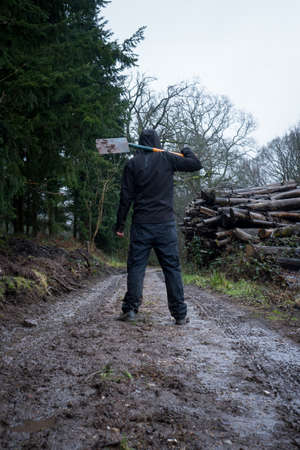 A hooded man, back to camera on a muddy path holding a spade, next to a log pile on a grey moody day...