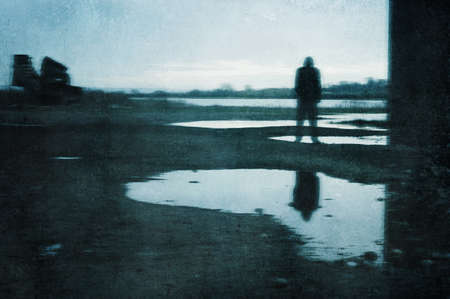 A moody figure, reflected in a puddle. Standing under a bridge in a post industrial landscape. With...