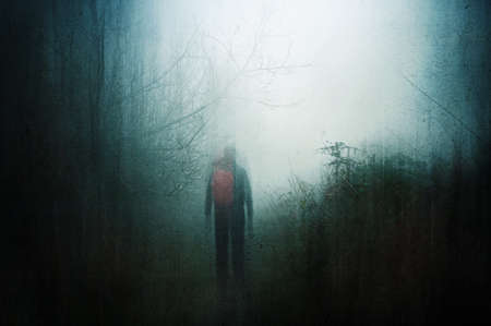 A horror concept of a hiker with a rucksack on a path through a spooky forest on a moody, foggy...