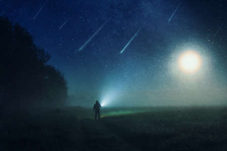 A man standing in the countryside shinning a torch into the darkness. With shooting stars heading towards the earth, against the night sky, With an abstract, artistic, editの写真素材