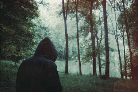 A spooky hooded figure, back to camera. Standing on the edge of a forest in the countryside. With a textured, grunge edit.の写真素材