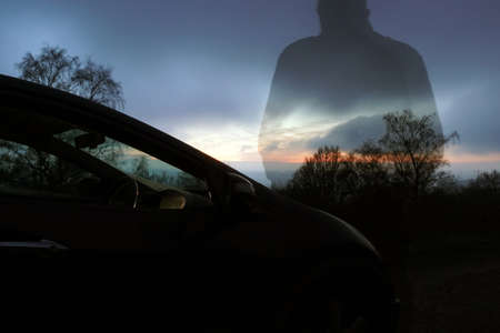A moody double exposure of a man next to a car looking out as the sun sets on a winters evening.の写真素材