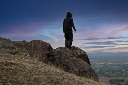 A moody, hooded man, back to camera standing on top of a mountain looking at a beautiful sunset.の写真素材