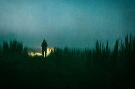 A dark scary concept. Of a mysterious figure, standing outside. Silhouetted against torch light at night. With a grunge, textured edit.の写真素材