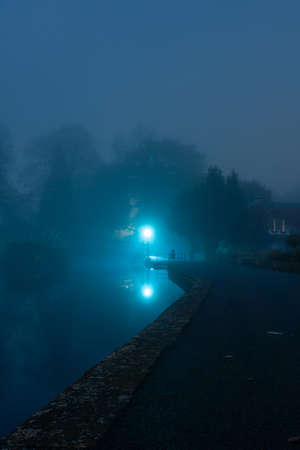 A spooky park with a lake, with trees silhouetted against lights on a path at night. With a moody edit.の写真素材