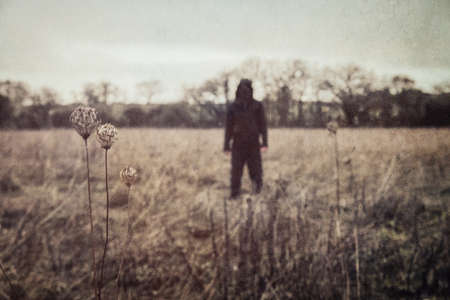 A horror concept of a hooded figure. Standing in the countryside. With a grunge, vintage edit