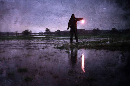 A mystery concept . Of a lost lonely hooded figure holding a lamp, standing in a flooded field at...