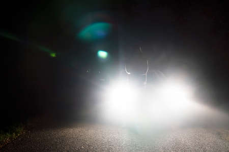 A sinister hooded figure. Standing in front of car headlights. On a moody night