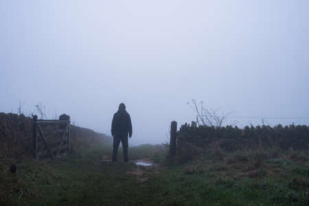 A mysterious hooded figure, back to camera. Standing on a track by a gate on a spooky, misty autumn day.の写真素材