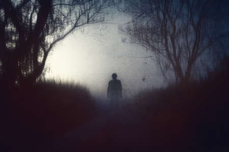 A spooky, horror concept. Of a scary shadow person with a hat standing in a country road.  With...