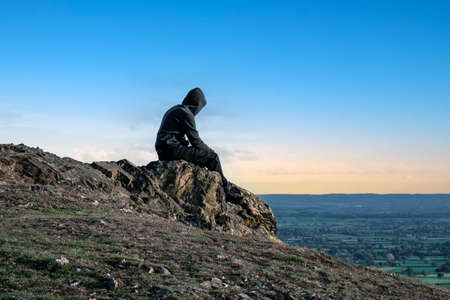 A mental health edit of a lonely hooded figure, looking out on the countryside on a summers evening.の写真素材