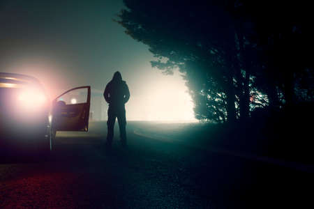 A dark moody concept of a threatening hooded figure standing next to a parked car. On a city street at nightの写真素材