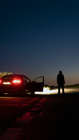 A mysterious hooded figure standing next to a parked car at night. On a spooky empty country road. With a vertical format.の写真素材