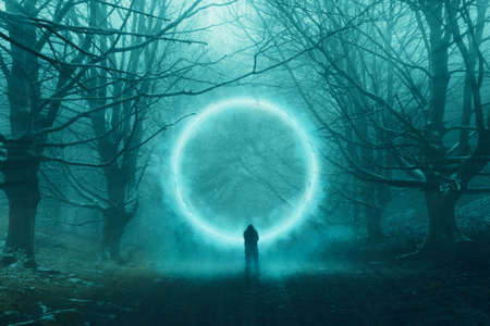 A spooky hooded figure, standing in front of a magical glowing portal floating in a forest. In a mysterious, foggy, winter forest.の写真素材
