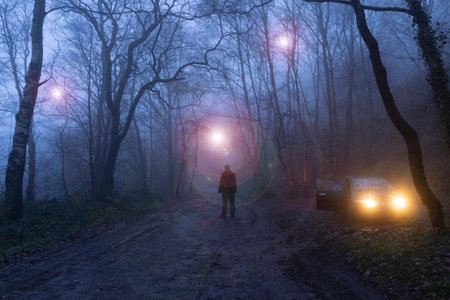 A man standing next to car watching glowing UFO's floating through trees. In a spooky forest on a foggy winters night.の写真素材