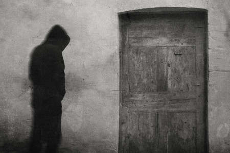 A ghostly spooky hooded monk. Standing by an old wooden door. With a grunge, vintage black and white edit.の写真素材