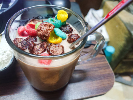Glass mug of chocolate milk topped with colorful cereal pieces and a metal spoon, resting on a...
