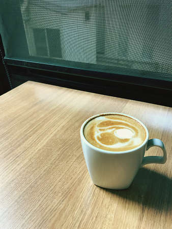 A hot latte in a white cup of coffee placed on a wooden table creates a warm morning atmosphere.の写真素材