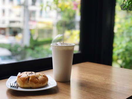 A bun with red bean paste topped with black sesame seeds is placed on a white plate, and there is a mug of white iced coffee on a brown wooden table in a cafe atmosphere.の写真素材