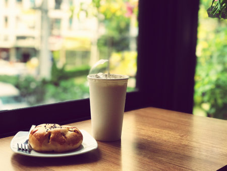 A bun with red bean paste topped with black sesame seeds is placed on a white plate, and there is a mug of white iced coffee on a brown wooden table in a cafe atmosphere.の写真素材