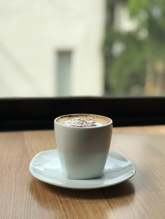A cup of coffee on a minimal wooden table placed by a large glass window overlooking the green...