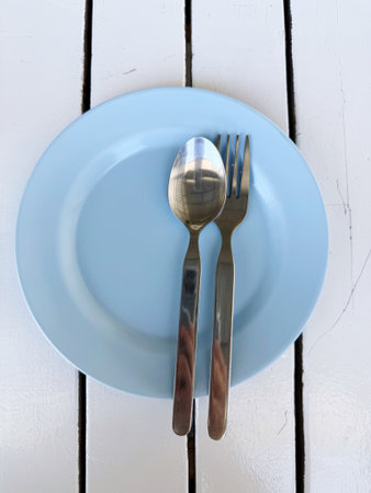 Empty blue plate with spoon and fork on white wooden table, top view
