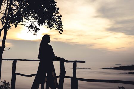 Trang, Thailand - 11 May 2018: Silhouette lady stand holding balcony in front of Hill view of ocean fog in dawn timeの写真素材