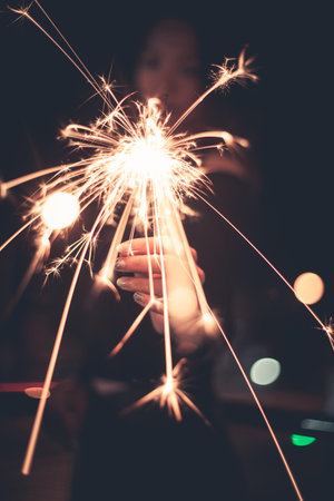 Lady holding sparklers stick on fire show forward, flame light spread out to camera at outdoor with light bokeh of light bulb at night timeの写真素材