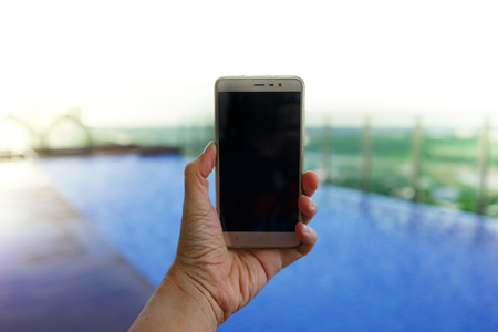 Hand holding mobile phone on swimming pool background. Closeup copy space photoの写真素材